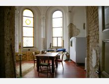 Ein Raum mit hohen, schmalen Bögen und bunten Glasfenstern, erkennbar durch Putzschäden an den Wänden und teilweise sichtbares Mauerwerk. Im Vordergrund steht ein einfacher Holztisch mit Stühlen, daneben ein Leuchter mit einer Kerze; auf der rechten Seite befinden sich ein weißer Paravent sowie diverse Möbelstücke und Gegenstände.