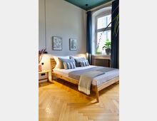 Ein Schlafzimmer mit einem schlichten Holzbett, dekoriert mit grauen und weißen Kissen sowie einer passenden Decke. Der Raum verfügt über einen Fischgrät-Holzboden, hohe Decken, ein großes Fenster mit dunkelblauen Vorhängen und Pflanzen auf der Fensterbank.