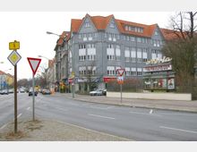 Straßenszene mit einem grauen, mehrstöckigen Gebäude mit Spitzdächern und orangefarbenen Ziegeln. Im Erdgeschoss befinden sich Geschäfte wie ein Copy-Shop und andere lokale Läden, während auf der Straßenseite Verkehrsschilder und eine Bushaltestelle mit Werbetafel sichtbar sind.