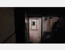 Ein dunkler, schmaler Kellerraum mit abblätternden Wänden und sichtbaren Rohren an der Decke. Im Hintergrund führen mehrere offene Türen in angrenzende, teilweise beleuchtete Räume.