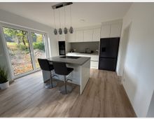 Moderne Küche mit weißer Einbauzeile, schwarzem Kühlschrank und einer zentralen Kücheninsel mit grauer Arbeitsplatte. Große Fenster lassen viel Tageslicht herein und bieten Blick auf eine Waldlandschaft; der Boden ist mit Holzoptik-Fliesen ausgelegt.