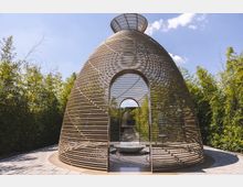 Moderne, kuppelförmige Metallstruktur aus horizontalen Streben mit einem rundbogigen Eingang, umgeben von dichtem Bambus und einem gepflasterten Weg; im Inneren befindet sich eine runde Sitzfläche.