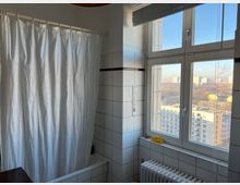 Badezimmer mit weißen Fliesen und einer Badewanne, die durch einen weißen Duschvorhang verdeckt ist. Rechts befinden sich ein großes Fenster mit Blick auf umliegende Wohngebäude und ein Heizkörper unter der Fensterbank.