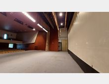 Bühneninnenraum eines Auditoriums mit Teppichboden, einer holzverkleideten Rückwand und seitlichen Vorhängen. Rechts ist eine helle Wand, und im Hintergrund sind Beleuchtungselemente und eine Tür zu sehen; links befinden sich die Zuschauerreihen.