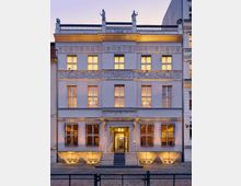 Fassade eines historischen Gebäudes mit klassizistischen Elementen, verziert mit Reliefs, Balkongeländer mit Statuen und symmetrisch angeordneten Fenstern. Der Eingang ist über eine kleine Treppe erreichbar, flankiert von vergitterten Fenstern, und das Gebäude ist stimmungsvoll beleuchtet.