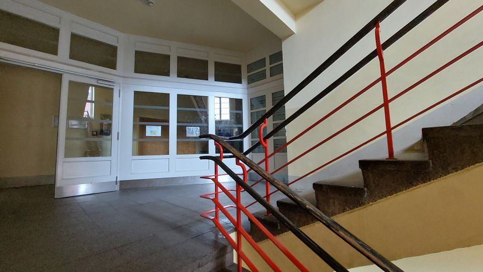 Treppenhaus in einem öffentlichen oder Wohngebäude mit einer Stein- oder Betontreppe, schwarzem Handlauf und roten Geländerstreben. Im Hintergrund sind weiße Türen mit großen Glaseinsätzen zu sehen, die einen weiteren Raum abschließen.
