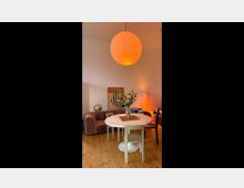 Wohnzimmer mit Holzdielenboden, einer weißen runden Tischgruppe und einem Sofa in Rosa. Eine große Papierlampe mit warmem Licht hängt von der Decke, während am Tisch Blumen in einer Vase stehen. Im Hintergrund sind eine Stehlampe, ein Gemälde und weitere Möbel erkennbar.