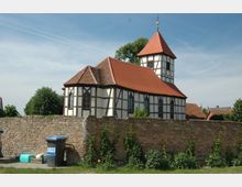 Fachwerkkirche mit roten Ziegeldächern und Turm, umgeben von einer niedrigen Backsteinmauer; im Vordergrund stehen Mülltonnen und einzelne Pflanzen. Der Himmel ist klar und blau, und im Hintergrund sind Bäume und Dächer weiterer Gebäude sichtbar.