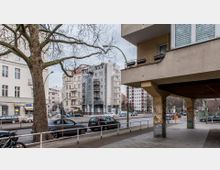 Straßenszene in einer Stadt mit mehrstöckigen Wohngebäuden unterschiedlicher Architektur, darunter Alt- und Neubauten. Im Vordergrund steht ein Baum, parkende Autos, ein überdachter Gehweg mit Graffiti an den Säulen und ein Geländer entlang des Gehwegs.