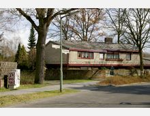 Wohnhaus aus der Nachkriegszeit mit hellbrauner Klinkerfassade und dunkelroten Fensterrahmen, umgeben von Bäumen und einem niedrigen Backsteinzaun. Straßenschilder an einer Straßenlaterne markieren die Kreuzung von "Am Sandwerder" und "Tilmansweg".