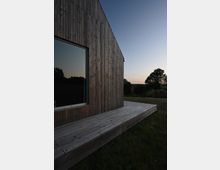 Holzhaus mit moderner Vertäfelung, Teilansicht der Fassade mit großem Fenster; davor eine einfache Holzterrasse. Umgeben von einer Wiese und vereinzelten Bäumen im Hintergrund bei Sonnenuntergang.