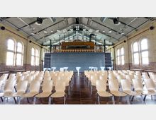 Ein Veranstaltungsraum mit Reihen aus hellen Holzstühlen, einer Bühne im Hintergrund sowie hohen Fenstern mit Bögen und Backsteinwänden. Die Decke ist offen mit sichtbaren Metallträgern und Beleuchtungselementen.