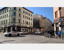 Straßenecke in einer Stadt mit mehrstöckigen, hell gestrichenen Gebäuden, darunter ein Burger-Restaurant und ein Sushi-Lokal im Erdgeschoss. Der Gehweg ist belebt, und im Vordergrund stehen mehrere Autos sowie geparkte Fahrräder neben einer Fußgängerampel.