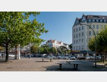Städtischer Platz mit gepflastertem Boden, umgeben von Bäumen und Mehrfamilienhäusern mit roten Dächern und heller Fassade. Im Vordergrund stehen Sitzbänke, während im Hintergrund Autos, Fahrräder und ein Restaurant sichtbar sind.