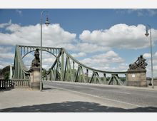 Grüne Stahlbogenbrücke mit kunstvollen Steinfiguren an beiden Seiten der Brückenpfeiler, typisches Straßenlaternen-Design, unter blauem Himmel mit vereinzelten Wolken. Die Brücke führt über einen Fluss und ist von Bäumen im Hintergrund umgeben.