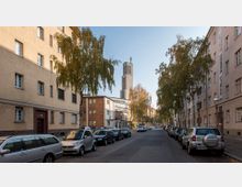 Straßenansicht mit parkenden Autos auf beiden Seiten, flankiert von Wohngebäuden aus der Vorkriegszeit. Im Hintergrund ist ein Kirchturm mit einer Uhr gut sichtbar, umgeben von Bäumen und herbstlichem Laub.