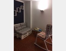 Ein kleiner, gemütlicher Raum mit einer Sitzbank aus Paletten mit grauen Polstern, einem weißen Schaukelstuhl mit Kissen, einem kleinen Tisch mit Büchern und Holzspielzeug sowie modernen weißen Wandregalen. Der Boden ist mit roten Fliesen ausgelegt, und die Wände sind teils weiß, teils dunkel gestrichen.