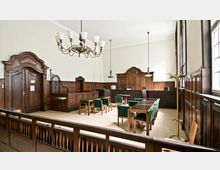 Ein Gerichtssaal mit holzvertäfelten Wänden, einer erhöhten Richterbank in der Mitte des Raumes, mehreren Holztischen und Stühlen mit grünen Polstern sowie einer großen Hängelampe aus Metall und Glas. Der Raum verfügt über hohe Decken, ein großes Fenster auf der rechten Seite und eine geschnitzte Holztür auf der linken Seite.