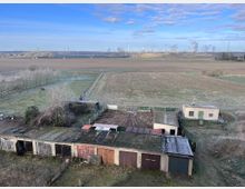 Ländliche Szene mit mehreren flachen, verwitterten Gebäuden, darunter Garagen mit verschiedenfarbigen Türen, und einem kleinen Nebengebäude. Im Hintergrund weite Felder, Baumreihen und Industrieanlagen mit Schornsteinen, aus denen Rauch aufsteigt.