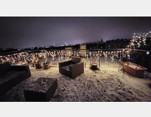 Dachterrasse bei Nacht mit leichtem Schneefall, dekoriert mit Lichterketten, mehreren Pflanzentöpfen und Sitzmöbeln aus Rattan. Im Hintergrund sind die Umrisse von Häusern und eine schwach beleuchtete Stadtlandschaft erkennbar.