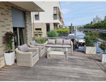 Moderne Terrasse mit Holzboden, möbliert mit einer Sitzgruppe aus geflochtenem Rattan mit grauen Kissen und einem Glastisch. Im Hintergrund befindet sich ein Kanal mit angrenzenden Wohngebäuden und Begrünung.