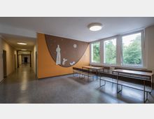 Ein heller Innenraum in einem Schul- oder Gemeindebereich mit großen Fenstern, die auf grüne Bäume blicken. An einer orangefarbenen Wand befindet sich ein dekoratives Kunstwerk mit einer Statue und Tiermotiven, daneben sind Tische und Bänke aufgestellt.