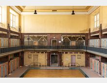 Innenraum eines historischen Schwimmbads mit gelben Fliesenwänden, einem leeren Pool mit Holzabdeckung und zwei Geschossen. Die oberen Etagen zeigen Fenster sowie Umkleidekabinen mit hellblauen Türen; Schriftzüge wie „Frauen: Duschen Toiletten“ und „Männer: Duschen Toiletten“ markieren Sanitärbereiche.