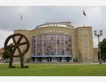 Außenansicht der Volksbühne in Berlin, einem historischen Theatergebäude mit einer hellen Steinfassade und einem auffälligen Schriftzug "VOLKSBÜHNE" über dem Eingang. Im Vordergrund steht eine große Kunstskulptur aus Metall in Form eines abstrakten Rads; die Szene ist von einer Wiese und einigen Bäumen umgeben.