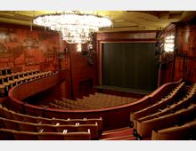 Innenansicht eines Theatersaals mit rötlich-brauner Holzverkleidung, einem großen Kristallleuchter an der Decke und Sitzreihen auf mehreren Ebenen. Eine Bühne mit geschlossenem Vorhang und Wandbilder mit kunstvollen Motiven sind sichtbar.