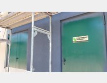 Eine metallische, grüne Tür mit einem Schild der Stadtwerke Schwedt GmbH (Station Niederkirchner Straße) an einer blauen Gebäudewand. Links im Bild befindet sich ein Gerüst, das teilweise die Ansicht verdeckt.