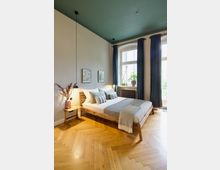 Schlafzimmer mit einem modernen Holzbett, dekoriert mit gestreiften Kissen und einer grauen Decke. Der Raum hat einen Fischgräten-Parkettboden, hohe Fenster mit dunklen Vorhängen und eine grüne Decke; auf dem Nachttisch steht eine Vase mit Zweigen, daneben hängt eine minimalistische Lampe.