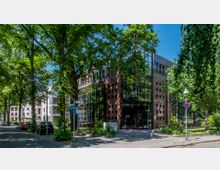 Moderne Bürogebäude mit Glasfassade, umgeben von Bäumen und Grünflächen, an einer ruhigen Straßenecke. Im Vordergrund sind Straßenschilder, geparkte Autos und ein Fußgängerkreuzungsbereich sichtbar.