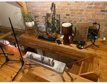 Holzraum mit offener Treppe, einer Backsteinwand und lose arrangierten Gegenständen wie einem Djembe-Trommel, Mikrofonständern, einer Topfpflanze und Kunstmaterialien. Der Boden besteht aus hellem Holz und ein Geländer aus Holzbalken grenzt den Bereich ab.