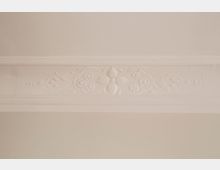 Detailaufnahme einer weißen Zimmerdecke mit breiter Stuckleiste und floralem Reliefmuster. Die glatte Deckenfläche wird von einer horizontal verlaufenden, verzierten Bordüre eingefasst.