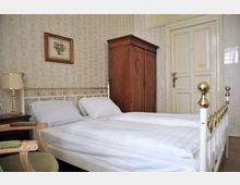 Schlafzimmer mit einem weißen Metallbett mit goldenen Akzenten und weißer Bettwäsche, einer antiken Holzkiste, einem cremefarbenen Wandschrank mit einer Tür und hell gemusterten Tapeten an den Wänden. Ein gerahmtes Blumenbild hängt über dem Bett.