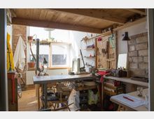 Eine helle, rustikale Werkstatt mit einem großen Arbeitsbereich aus Holz, umgeben von Werkzeugen an einer Sperrholz-Wand. Das Zimmer hat freiliegende Backsteinwände, eine Holzdecke und ein großes Fenster, das Tageslicht hereinlässt; im Raum befinden sich verschiedene handwerkliche Materialien und Pflanzen.