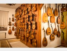 Raum mit einer Sammlung von Musikinstrumenten, hauptsächlich Gitarren und Lauten, die an Wänden auf Haken aufgehängt sind. Die Wände sind mit Holzelementen und dekorativem Stoff verkleidet, ergänzt durch klassische Inneneinrichtung mit Teppichen und Kronleuchtern.