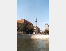 Blick auf die Spree mit dem Berliner Fernsehturm im Hintergrund, flankiert von Gebäuden mit historischer und moderner Architektur sowie Bäumen am Ufer. Der Himmel ist klar und blau.
