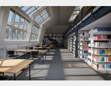 Lesehalle einer Bibliothek mit großen, schrägen Fenstern auf der linken Seite, die viel Tageslicht hereinlassen, und Regalen voller Bücher auf der rechten Seite. In der Mitte befinden sich Reihen von Holzschreibtischen mit schwarzen Stühlen, und im Hintergrund ist ein Treppenaufgang sichtbar.