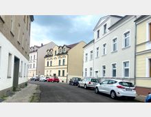 Straßenansicht eines Wohnviertels mit mehrstöckigen hellen Häusern in einfacher historischer Architektur. Entlang der Straße sind Autos geparkt, und der Himmel ist bewölkt.