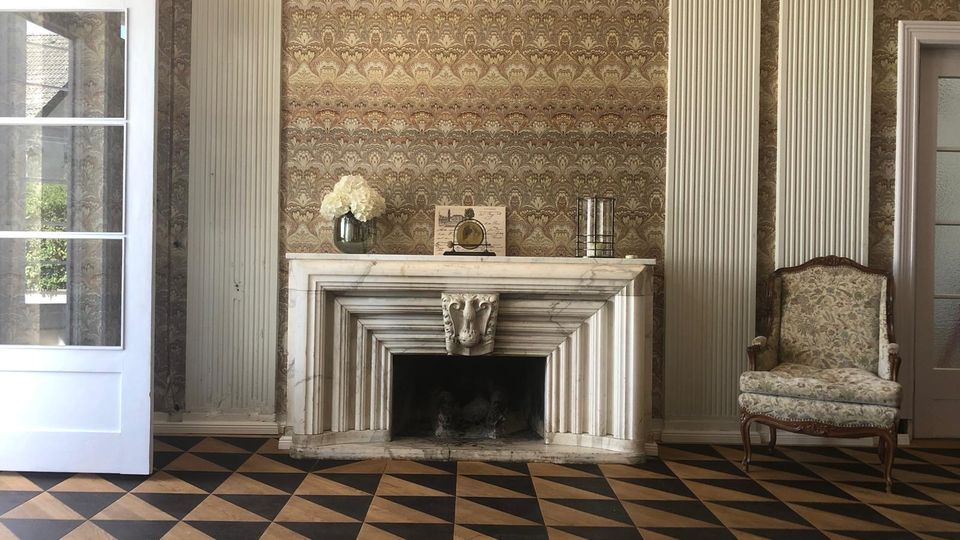 Klassischer Innenraum mit weißem Marmorkamin in der Mitte, gerahmt von hohen, geriffelten Wandpfeilern und einer gemusterten Tapete. Davor liegt ein schwarz-beiges Rautenkachelmuster am Boden; rechts steht ein gepolsterter Sessel, links ist eine geöffnete Glastür zu sehen.