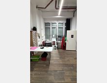 Small studio workspace with a wood floor, a folding table, storage drawers, and sewing equipment, facing a wall of tall windows with shutters. Exposed ceiling pipes and a fluorescent light hang overhead, with costume racks and a white door along the right wall.