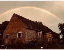 Backsteinbauernhaus mit steilem Ziegeldach und zwei kleinen Fenstern, umgeben von Wiesen und Bäumen. Im Hintergrund wölbt sich ein Regenbogen über den bewölkten Himmel.