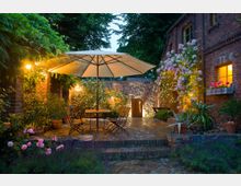 Gemütlicher Innenhof mit Kopfsteinpflaster, einer zentralen Sitzgruppe mit Sonnenschirm und umgeben von Blumenbeeten mit Rosen und Lavendel. Die Umgebung umfasst eine Backsteinwand, ein bewachsenes Haus und warme Beleuchtung, die eine einladende Atmosphäre schafft.
