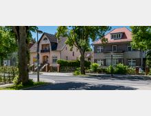 Straßenszene in einem grünen Wohnviertel mit zwei freistehenden Häusern: links ein helles Einfamilienhaus mit steilem Giebeldach und Balkon, rechts ein dunkles Backsteinhaus mit rotem Dach und großen Fenstern. Im Vordergrund verlaufen Straße und Gehweg, eingerahmt von hohen Bäumen, Hecken und niedrigen Zäunen.