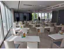 Konferenzraum mit hellen, modernen Möbeln, bestehend aus weißen Tischen und Stühlen, angeordnet in Reihen. Links befinden sich bodentiefe Fenster, die viel Tageslicht einlassen, während im Hintergrund große Pflanzen und ein Rednerpult sichtbar sind.