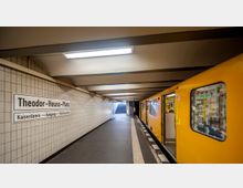 U-Bahnhof mit gelbem Zug an einem Bahnsteig, gefliesten Wänden und einem Schild mit der Aufschrift "Theodor-Heuss-Platz". Eine Treppe am Ende des Bahnsteigs führt nach oben.