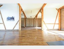 Ein heller, offener Raum mit Holzboden, freiliegenden Holzbalken und teilweise weiß gestrichenen sowie unverputzten Ziegelwänden. Große Fenster lassen viel Tageslicht herein, und an einer Wand hängt ein gerahmtes Bild.