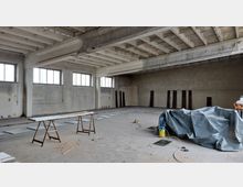 Großer, leerer Innenraum einer Halle mit roh belassenen Betonwänden und einer Betondecke mit sichtbaren Querträgern. Im Vordergrund liegen Arbeitsmaterialien wie Holzgestelle, Plastikfolie und Werkzeuge; an den Wänden befinden sich rechteckige Fenster.