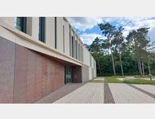 Modernes Rathausgebäude in Dallgow-Döberitz mit einer Fassade aus roten und weißen Paneelen, großen schmalen Fenstern und einer Glas-Eingangstür. Im Hintergrund sind hohe Bäume und Grünflächen zu sehen.